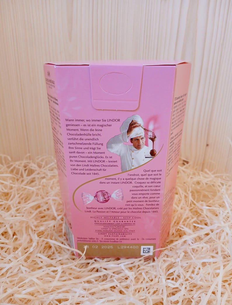 May include: A pink and white box containing a chocolate truffle. The box features an image of a chef holding a whisk and a bowl of chocolate. The text on the box reads "Wann immer, wo immer Sie LINDOR geniessen - es ist ein magischer Moment. Wenn die feine Chocoladenh&uuml;lle bricht, verf&uuml;hrt die unendlich zartschmelzende F&uuml;llung Ihre Sinne und tr&auml;gt Sie sanft davon - ein Moment  puren Chocoladengl&uuml;cks. Es ist Ihr Moment. Mit LINDOR - kreiert von den Lindt Ma&icirc;tres Chocolatiers. Liebe und Leidenschaft f&uuml;r Chocolade seit 1845." The text on the bottom of the box reads "HIGHLY MELTABLE KEEP COOL QUALITY GUARANTEE MANUFACTURED BY LINDT & SPRUNGLI AG LINDT & SPRUNGLI (UK) LTD LINDT SUSTAINABILITY 02 2025 L294400"