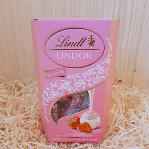 May include: Pink box of Lindt Lindor chocolates with a strawberry and cream flavour. The box features a white chocolate truffle with a strawberry on top and a pink background with white floral designs. The text on the box reads "Lindt Lindor Fraise Erdbeere Unendlich Zartschmelzend Fraise & Creme Erdbeere & Rahm."