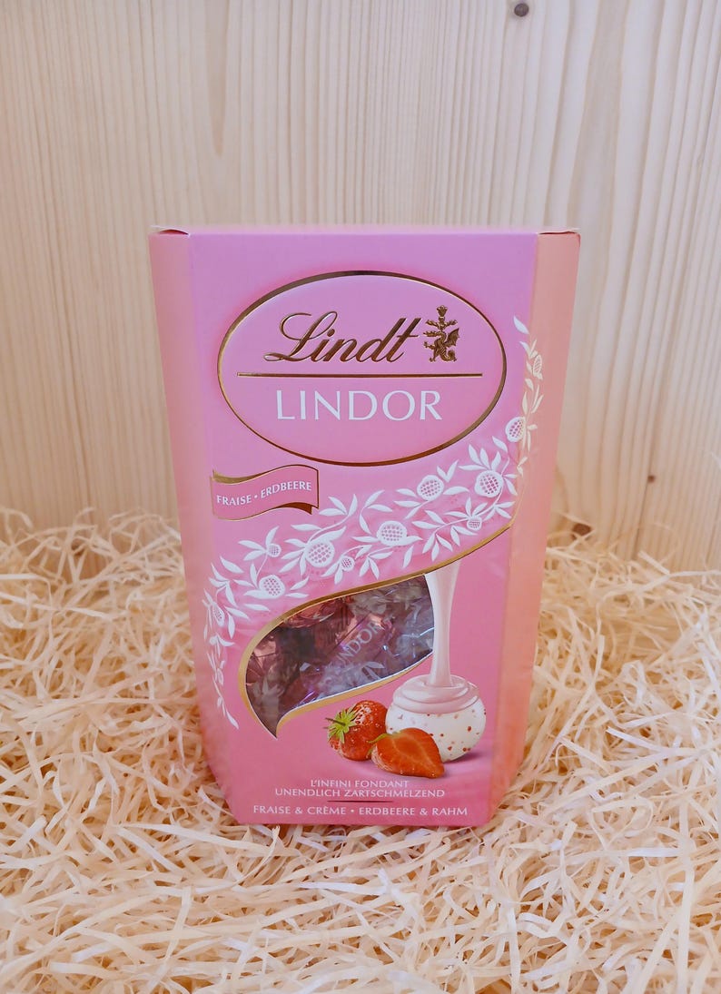 May include: Pink box of Lindt Lindor chocolates with a strawberry and cream flavour. The box features a white chocolate truffle with a strawberry on top and a pink background with white floral designs. The text on the box reads "Lindt Lindor Fraise Erdbeere Unendlich Zartschmelzend Fraise & Creme Erdbeere & Rahm."