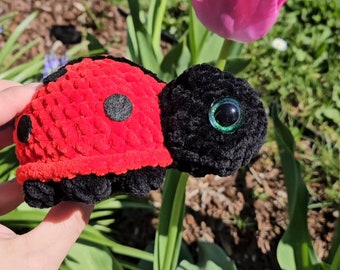 May include: A crocheted ladybug toy with black and red yarn, and large green eyes. The ladybug is sitting on a green plant with pink flowers.
