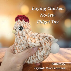 May include: A crocheted laying chicken toy with a red comb and a white egg. The toy is brown and white with a fuzzy texture. The text "Laying Chicken No-Sew Fidget Toy" is above the toy. The text "Pattern by Crystals Cute Creationz" is below the toy.