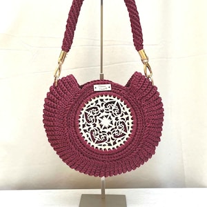 May include: A burgundy crochet handbag with a circular design and a white, intricate, laser-cut centerpiece. The bag features a braided handle and gold-coloured hardware. The bag is displayed on a metal stand.