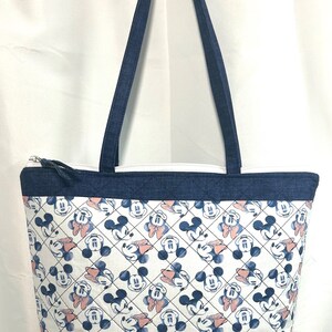 May include: A quilted tote bag featuring a repeating pattern of Mickey and Minnie Mouse heads on a white background with a diamond grid overlay. The bag has dark blue denim-like fabric at the top and bottom edges, matching shoulder straps, and a zipper.