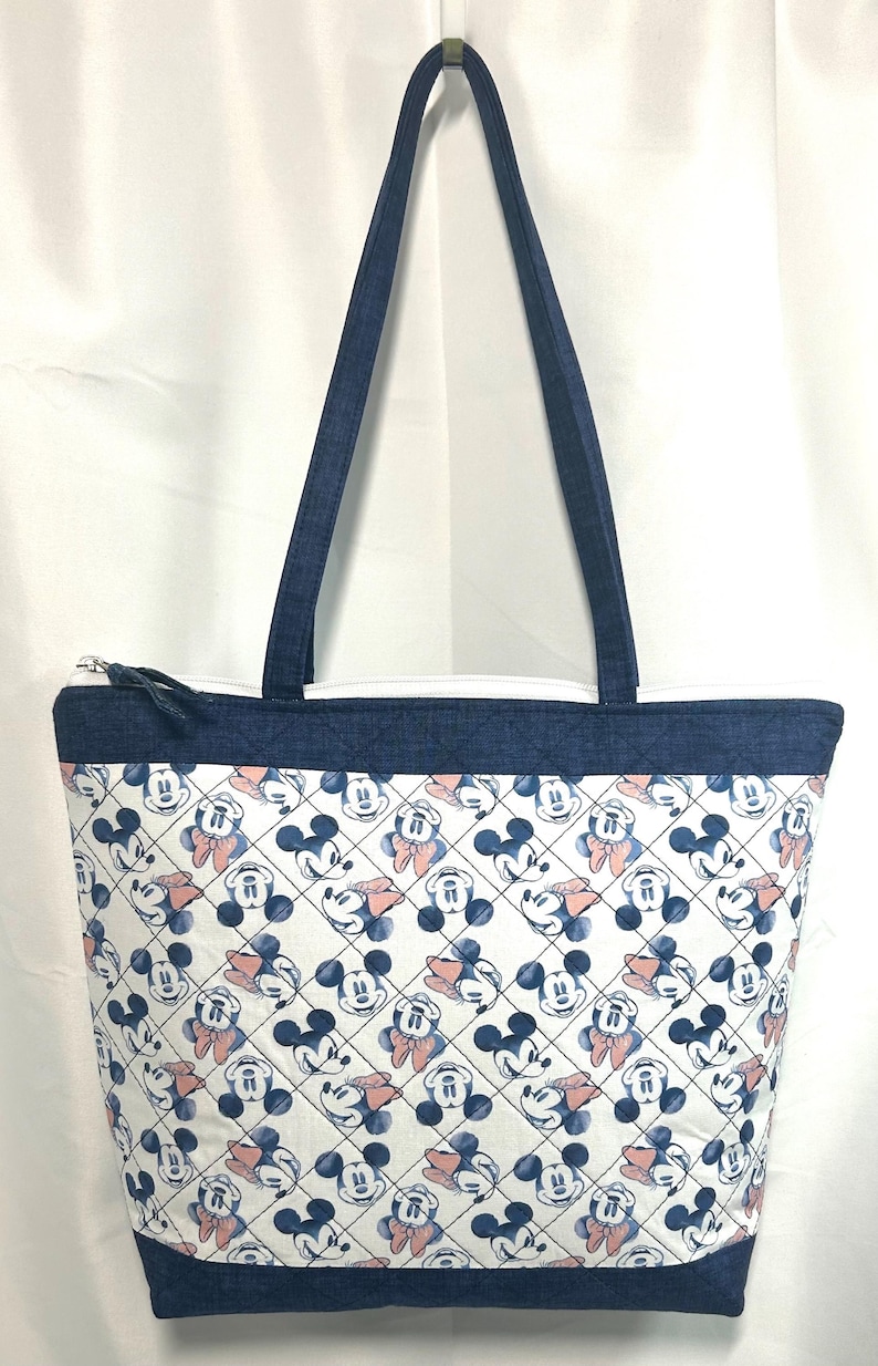 May include: A quilted tote bag featuring a repeating pattern of Mickey and Minnie Mouse heads on a white background with a diamond grid overlay. The bag has dark blue denim-like fabric at the top and bottom edges, matching shoulder straps, and a zipper.