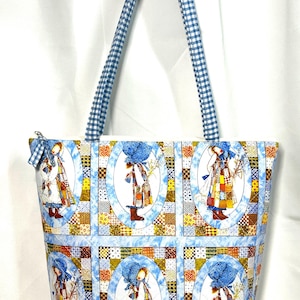 May include: A tote bag featuring a repeating pattern of figures in blue bonnets and patchwork dresses within oval frames. The fabric includes blue and white gingham handles and a zipper closure with a small matching gingham fabric pull tab.