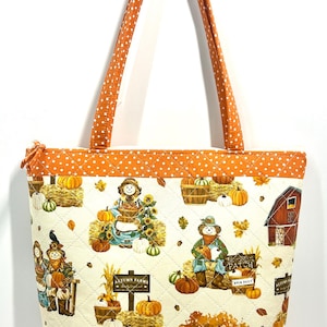 May include: A quilted tote bag featuring an off-white fabric with a repeating autumn-themed print of scarecrows, pumpkins, hay bales, and a red barn. The bag has orange polka-dot fabric trim on the top and bottom edges and two matching straps.