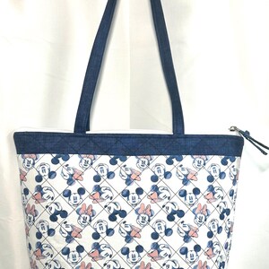 May include: A tote bag featuring a repeating pattern of Mickey and Minnie Mouse faces in blue and soft pink on a white background. The bag has dark blue denim-textured fabric at the top and bottom with a quilted diamond stitch pattern and handles.