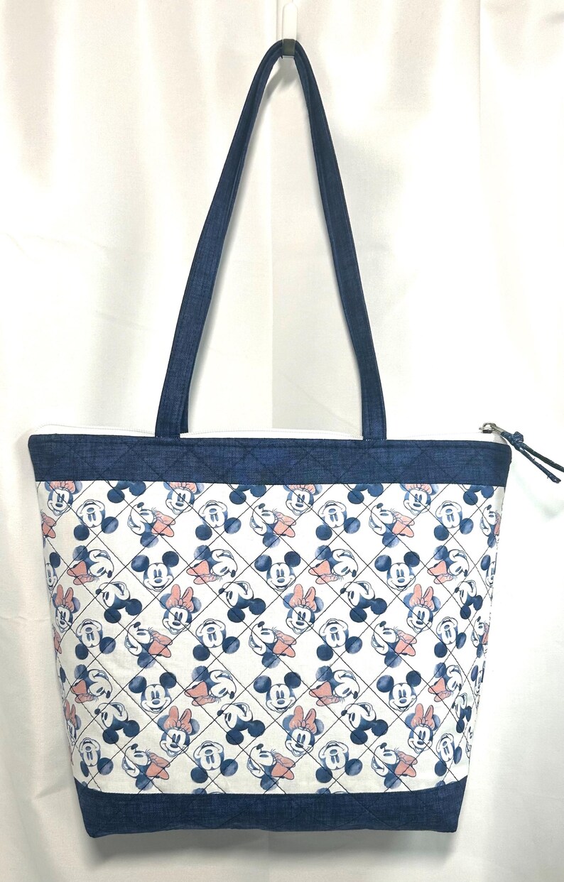 May include: A tote bag featuring a repeating pattern of Mickey and Minnie Mouse faces in blue and soft pink on a white background. The bag has dark blue denim-textured fabric at the top and bottom with a quilted diamond stitch pattern and handles.