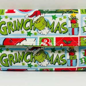 May include: A quilted fabric clutch featuring a repeating pattern of the Grinch, the text "GRINCHMAS," and "12 DAYS OF," along with illustrations of gift boxes and red Santa hats on a white background with green stars and blue accents.