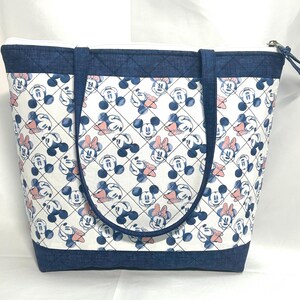 May include: A tote bag featuring a white fabric body with a repeating pattern of blue Mickey Mouse and pink Minnie Mouse heads inside a black diamond grid. The bag has dark blue denim-textured trim at the top and bottom, two handles, and a zipper.