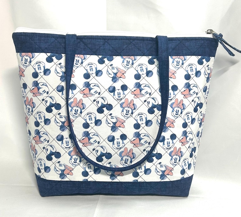May include: A tote bag featuring a white fabric body with a repeating pattern of blue Mickey Mouse and pink Minnie Mouse heads inside a black diamond grid. The bag has dark blue denim-textured trim at the top and bottom, two handles, and a zipper.