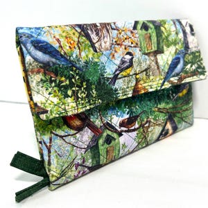 May include: A rectangular quilted fabric clutch featuring a colorful print of various birds, including bluebirds and chickadees, perched on branches near wooden birdhouses amidst green foliage and trees. The clutch has a fold-over flap and dark green tabs.