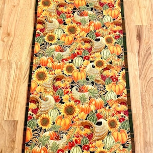 Puede incluir: Camino de mesa rectangular con temática de cosecha de otoño. El corredor presenta un patrón repetido de calabazas, girasoles, cuernos de la abundancia y calabazas en tonos naranja, amarillo y verde sobre un fondo negro. El borde es un patrón de cuadros.