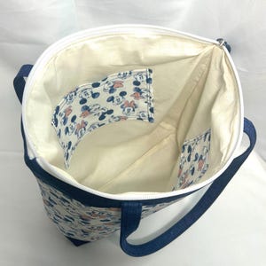 May include: A fabric tote bag with a white zipper closure and dark blue straps. The interior is lined with cream-colored fabric and features two internal pockets decorated with a repeating pattern of Mickey and Minnie Mouse faces.