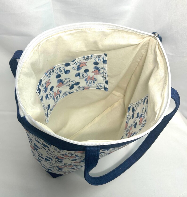 May include: A fabric tote bag with a white zipper closure and dark blue straps. The interior is lined with cream-colored fabric and features two internal pockets decorated with a repeating pattern of Mickey and Minnie Mouse faces.