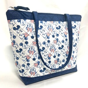 May include: A quilted tote bag featuring a white fabric with a repeating pattern of blue Mickey Mouse and Minnie Mouse faces, accented by peach-colored bows. The bag includes dark blue denim-textured trim, handles, and a top zipper closure.