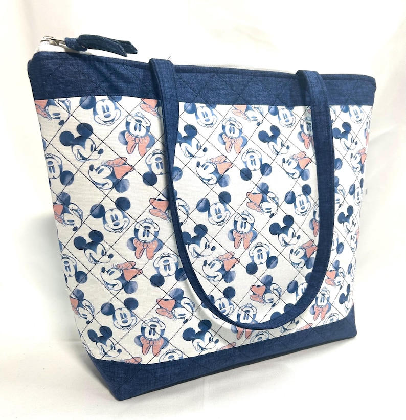 May include: A quilted tote bag featuring a white fabric with a repeating pattern of blue Mickey Mouse and Minnie Mouse faces, accented by peach-colored bows. The bag includes dark blue denim-textured trim, handles, and a top zipper closure.