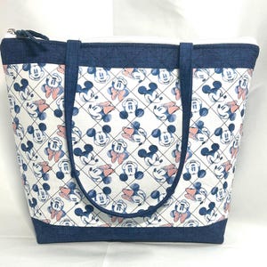 May include: A quilted tote bag featuring a repeating pattern of Mickey and Minnie Mouse faces on a white background. The bag has dark blue denim-textured fabric trim at the top and bottom, matching dark blue straps, and a top zipper closure.