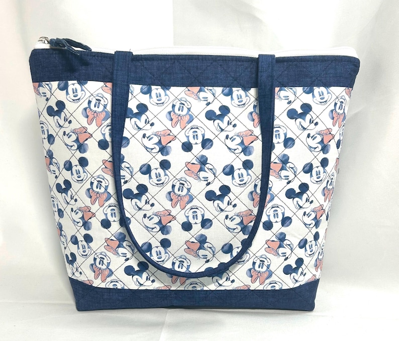 May include: A quilted tote bag featuring a repeating pattern of Mickey and Minnie Mouse faces on a white background. The bag has dark blue denim-textured fabric trim at the top and bottom, matching dark blue straps, and a top zipper closure.