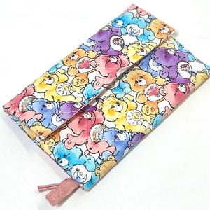 May include: A rectangular quilted fabric pouch featuring a colorful all-over print of various Care Bears characters in shades of blue, yellow, pink, and purple. The pouch has a fold-over flap closure and a small pink fabric pull tab at the bottom corner.