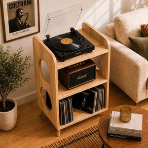 May include: A light-toned wooden record player stand featuring three open shelves, circular side cutouts, and short legs. The top shelf holds a black turntable, the middle shelf contains a rectangular brown speaker, and the bottom shelf stores vinyl records.