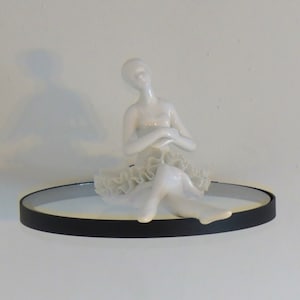 May include: A white porcelain figurine of a seated dancer with a ruffled skirt, positioned on a circular mirror base with a thin black frame. The dancer has arms crossed over the chest and legs extended forward, featuring a smooth, glossy finish.