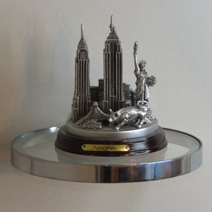 May include: A silver-toned metal New York City souvenir figurine featuring the Chrysler Building, Empire State Building, Statue of Liberty, and a charging bull. The sculpture sits on a circular dark brown wooden base with a gold-colored plaque.