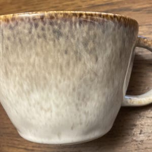 May include: A ceramic mug featuring a speckled, mottled glaze in shades of cream, beige, and brown. The rim and the handle are accented with a darker, concentrated brown tone that fades into the lighter, textured body of the cup.
