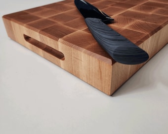 May include: A wooden cutting board with a checkered pattern and a black knife resting on top. The board has a handle on the side.
