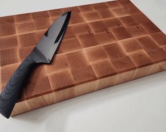May include: A rectangular end-grain wooden cutting board with a grid pattern of light and medium brown wood blocks. A kitchen knife with a black handle and a dark metallic blade rests diagonally across the surface of the wooden board.