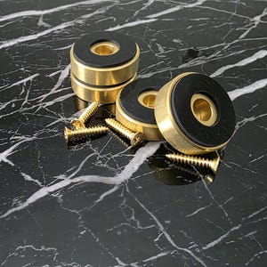 Op de afbeelding: Een verzameling goudkleurige, cirkelvormige hardwarecomponenten met zwarte rubberen inzetstukken. Verschillende gouden schroeven liggen in de buurt verspreid. De items zijn gerangschikt op een zwart, marmerachtig oppervlak.