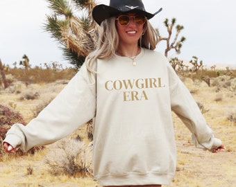 May include: A beige sweatshirt with the text "COWGIRL ERA" printed in gold on the front. The model is wearing a black cowboy hat and sunglasses.