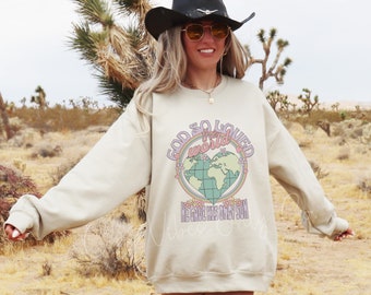 Puede incluir: Una sudadera beige con un estampado gráfico de un globo terráqueo en forma de corazón con un arcoíris y el texto "Dios amó tanto al mundo que dio a su Hijo único".