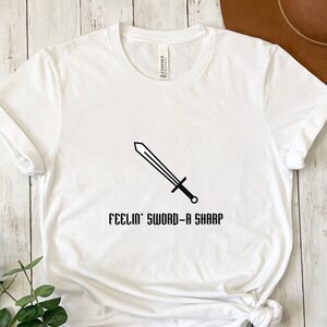 Pode incluir: Camiseta branca com um gráfico de espada preta e o texto "Feelin' Sword-a Sharp".