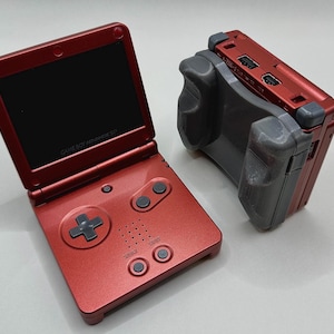 May include: A red Game Boy Advance SP handheld gaming console with a black screen and buttons. The console is closed and has a black and gray controller attachment.