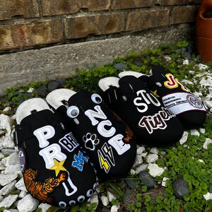 May include: Three black fabric clogs with various colorful patches and embroidered text. The clogs have a white strap and a white sole. The text on the clogs includes "Prince", "Houston Independent School District", and "Tiger".