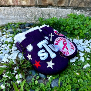 May include: Purple fuzzy slippers with a white fur lining. The slippers have various patches including a star, a red star, a cross, and a pink ribbon with the words "Believe, Fight, Hope".