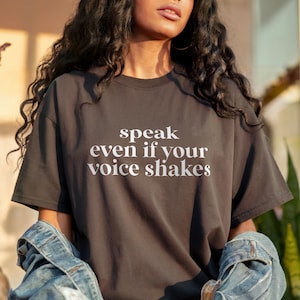 Op de afbeelding: Donkergrijs t-shirt met de witte tekst "speak even if your voice shakes". Het shirt heeft korte mouwen en een ronde hals. Het model draagt een spijkerjasje.