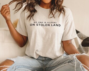 Könnte beinhalten: Eine Person trägt ein graues T-Shirt mit dem Aufdruck "NO ONE IS ILLEGAL ON STOLEN LAND".