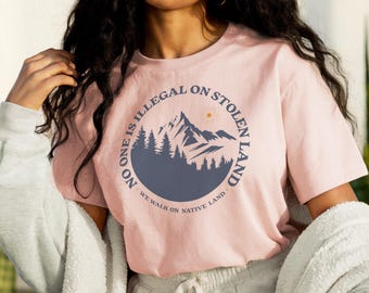 Może przedstawiać: Różowy T-shirt z grafiką przedstawiającą pasmo górskie i tekstem "No one is illegal on stolen land. We walk on native land."