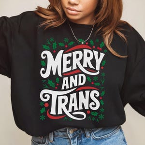 Puede incluir: Sudadera negra con el texto blanco "MERRY AND TRANS", detalles rojos y diseños de acebo y copos de nieve. Una prenda festiva.