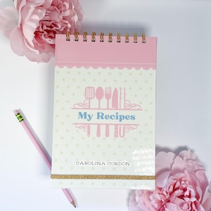 Puede incluir: Una libreta de espiral rosa y blanca con una espiral dorada. La portada tiene un fondo blanco con lunares rosas y una ilustración rosa y azul de utensilios de cocina. El texto "My Recipes" está en azul y el texto "CAROLINA CORDÓN" está en negro.