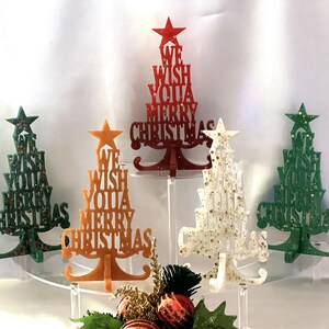 May include: Five decorative Christmas tree ornaments in various colors, including red, green, white, and orange. Each tree is made of wood and features the words "We Wish You a Merry Christmas" in a layered design. The trees are displayed on clear acrylic stands.