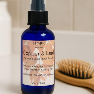 May include: A blue glass bottle of Copper & Leaf Ayurvedic Leave in Hair Serum with a black spray nozzle. The label includes the text "HOPE skincare" and product details. A wooden hairbrush is in the background.