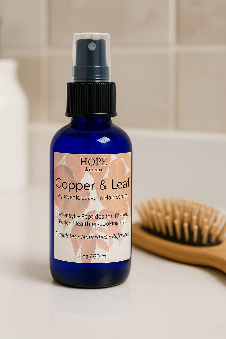 May include: A blue glass bottle of Copper & Leaf Ayurvedic Leave in Hair Serum with a black spray nozzle. The label includes the text "HOPE skincare" and product details. A wooden hairbrush is in the background.