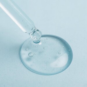May include: Close-up of a clear glass dropper dispensing a drop of transparent liquid onto a light blue surface. The liquid forms a large, round droplet with small bubbles, suggesting a serum or cosmetic product.