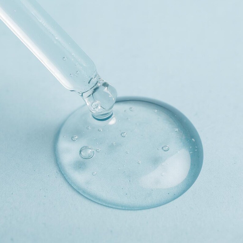 May include: Close-up of a clear glass dropper dispensing a drop of transparent liquid onto a light blue surface. The liquid forms a large, round droplet with small bubbles, suggesting a serum or cosmetic product.