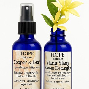 May include: Two blue glass bottles of HOPE skincare products. One is Copper & Leaf Ayurvedic Leave in Hair Serum, the other is Ylang Ylang Bloom Detangler. Each bottle is 2 fl oz / 60 ml. Yellow flowers and green leaves are in the Ylang Ylang bottle.