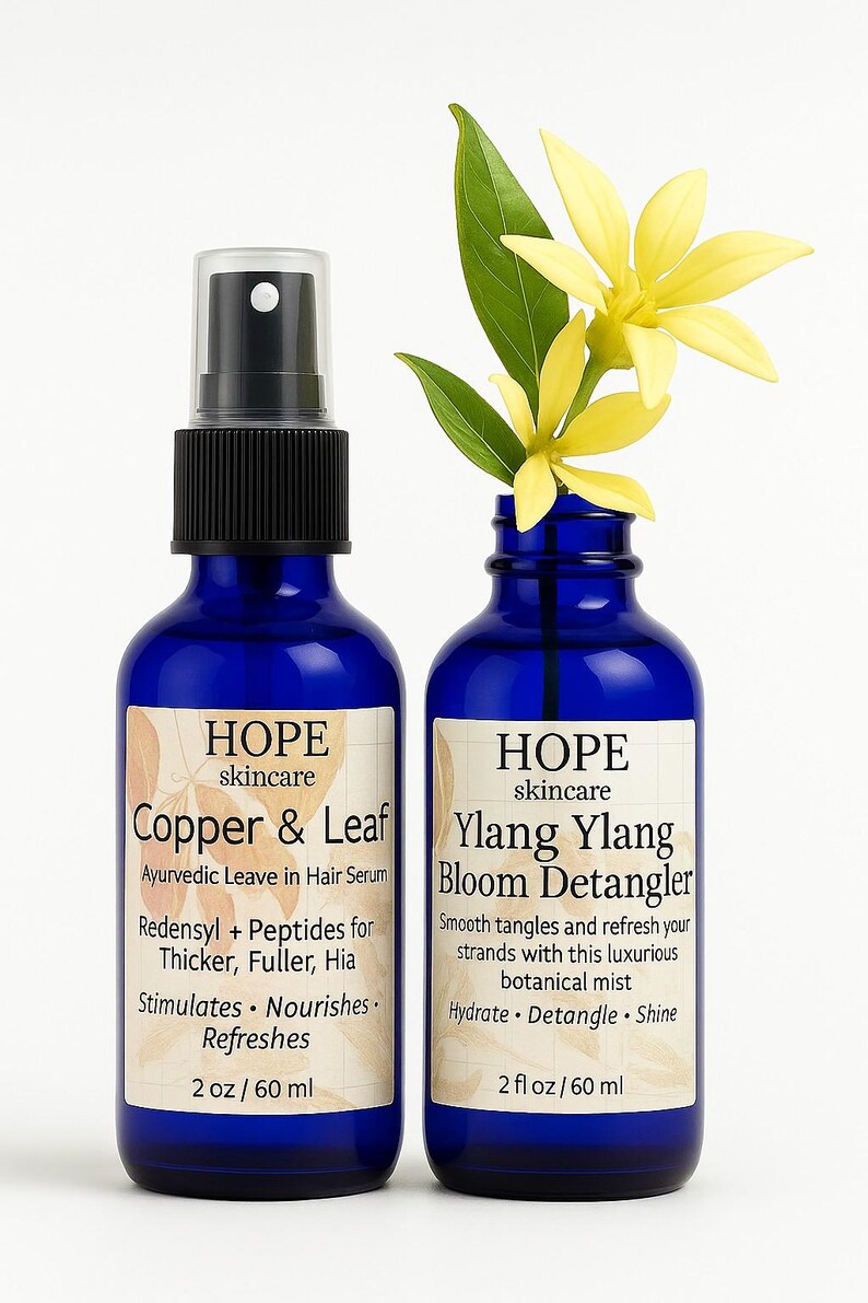 May include: Two blue glass bottles of HOPE skincare products. One is Copper & Leaf Ayurvedic Leave in Hair Serum, the other is Ylang Ylang Bloom Detangler. Each bottle is 2 fl oz / 60 ml. Yellow flowers and green leaves are in the Ylang Ylang bottle.