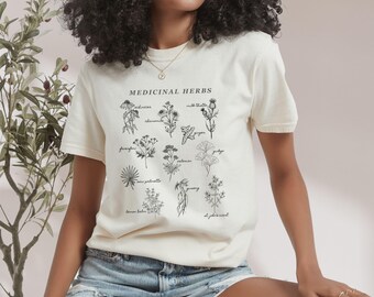 May include: A cream-colored t-shirt with a black graphic of medicinal herbs and their names. The graphic includes illustrations of milk thistle, echinacea, valerian, and more.